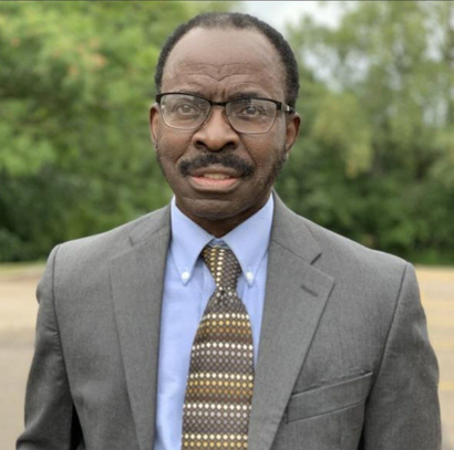 Man wearing glasses, suit, and tie, standing outside, looking forward.