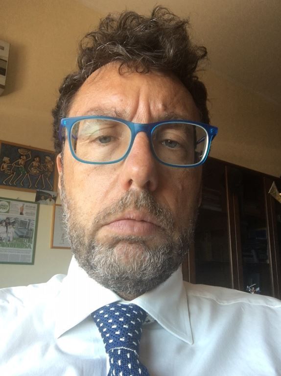 Man with curly brown hair, blue glasses, and a somber expression, wearing a white shirt and patterned tie.