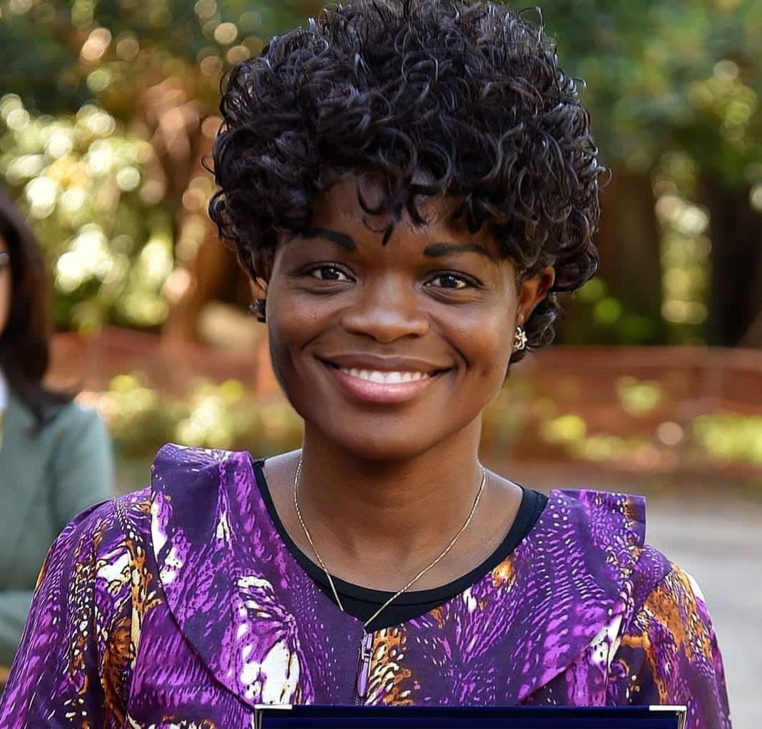 Man in blue suit, sunglasses, and patterned turban. Holds event badge. Pink and purple background.