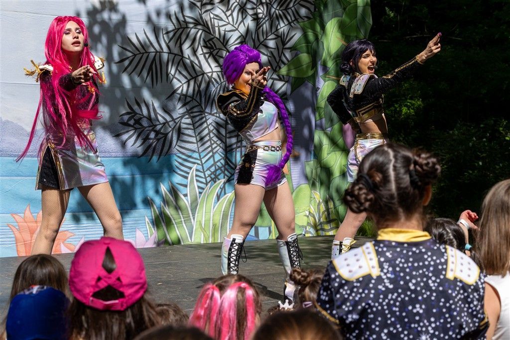 Three animated pop singers in colorful outfits hold microphones and pose against a sparkling blue stage background.