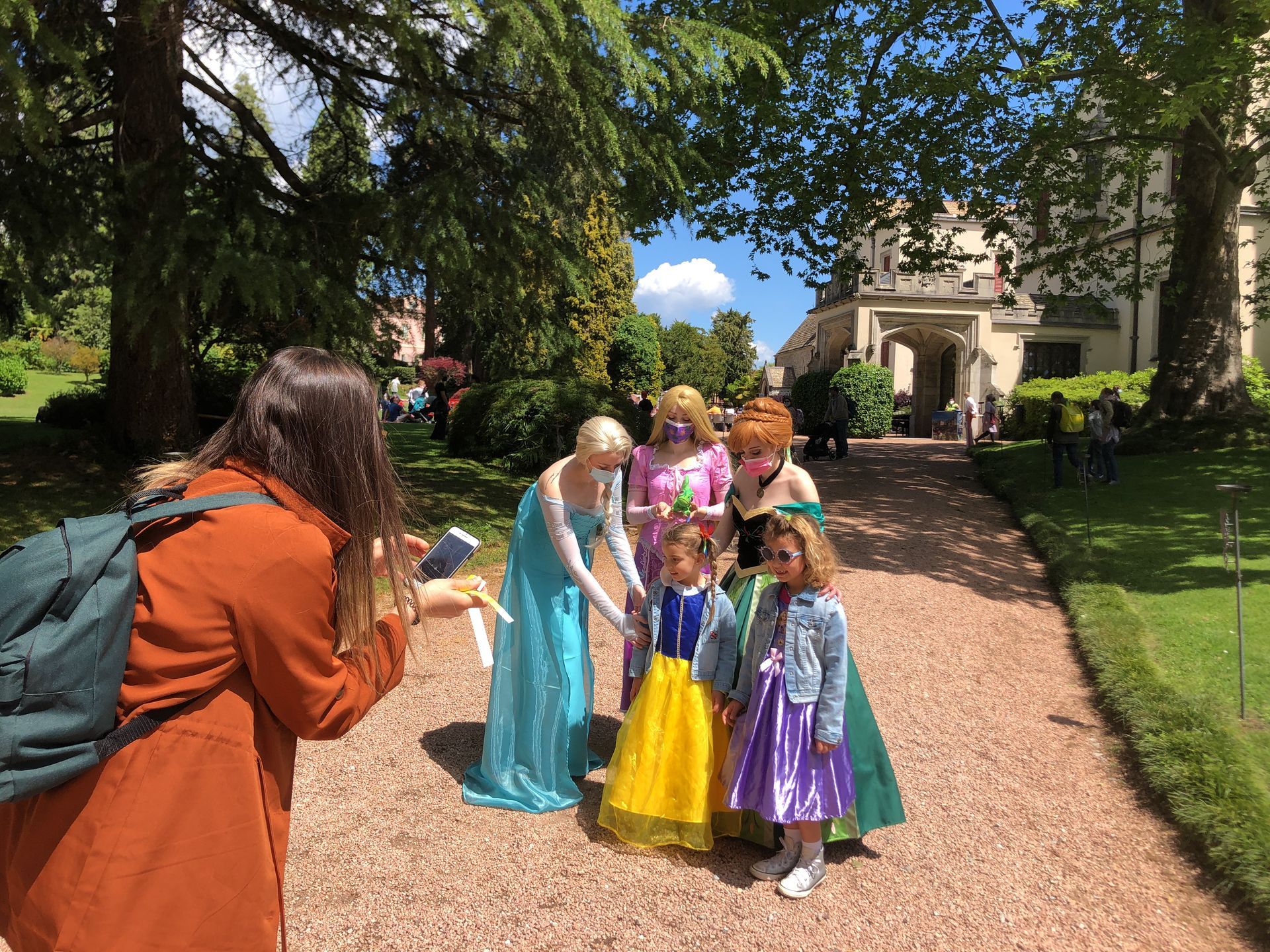 Una persona con una giacca arancione fotografa due bambine vestite da principesse e tre personaggi in costume in un giardino.