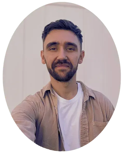 Man with a beard wearing a beige shirt and white t-shirt smiles, standing in front of a plain wall.