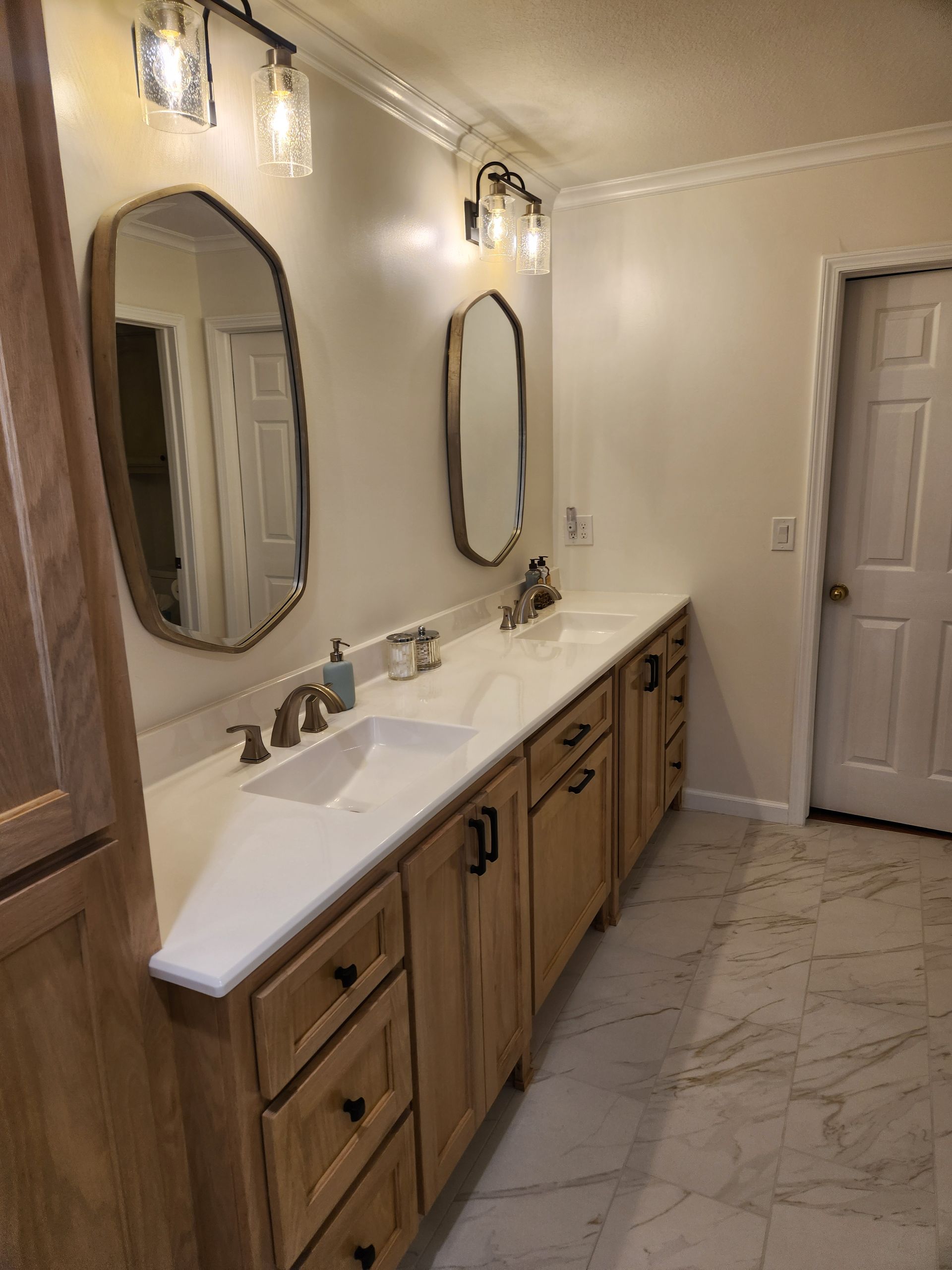 Bathroom with light wood cabinets, two sinks, mirrors, and a white countertop. Beige walls and marble floors.