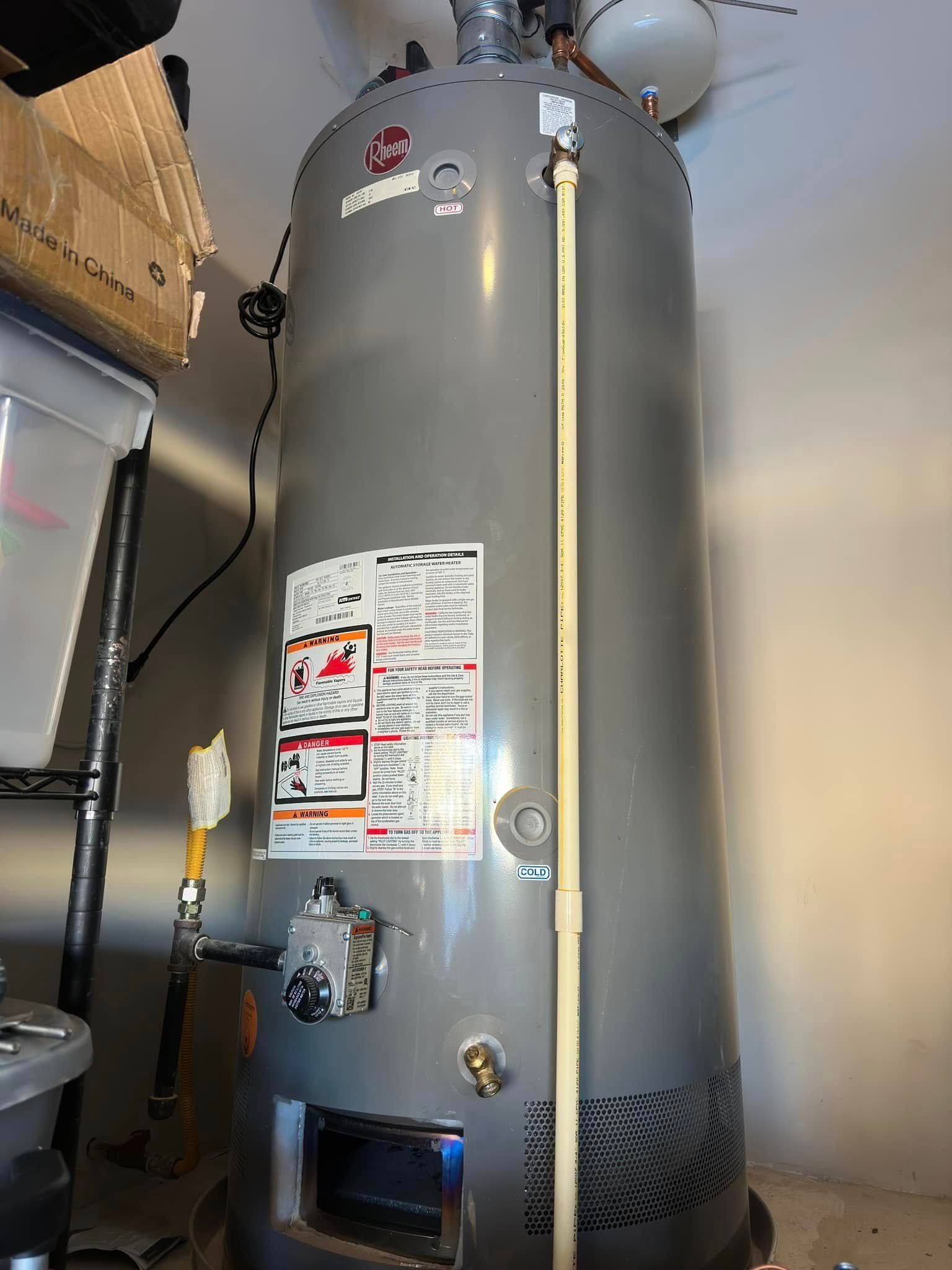 A tall, grey water heater in a basement.  A white plastic pipe runs along the side.  A gas line connects on the left.