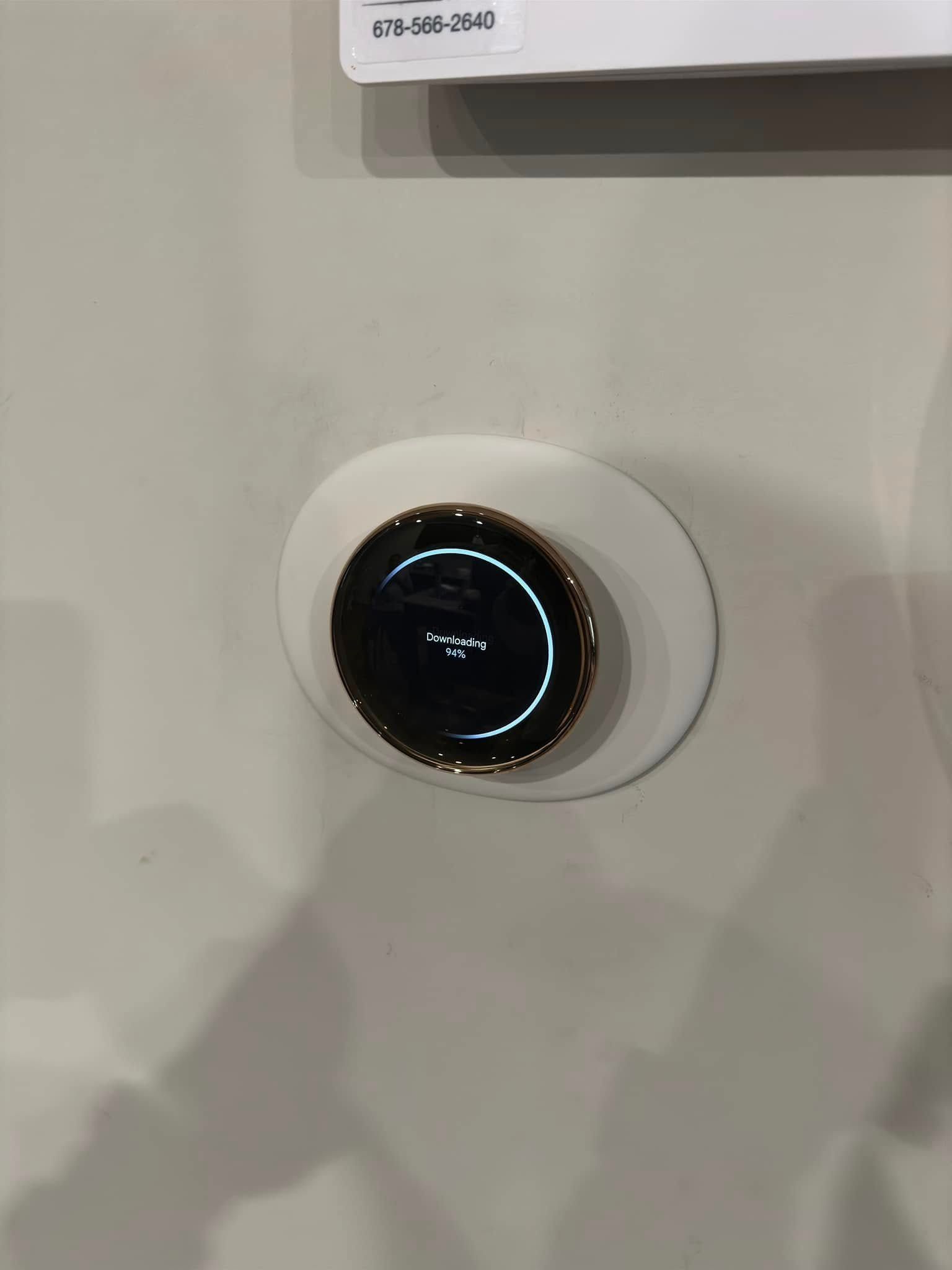 Round, black and gold thermostat on a white, oval-shaped base, mounted on a grey wall.