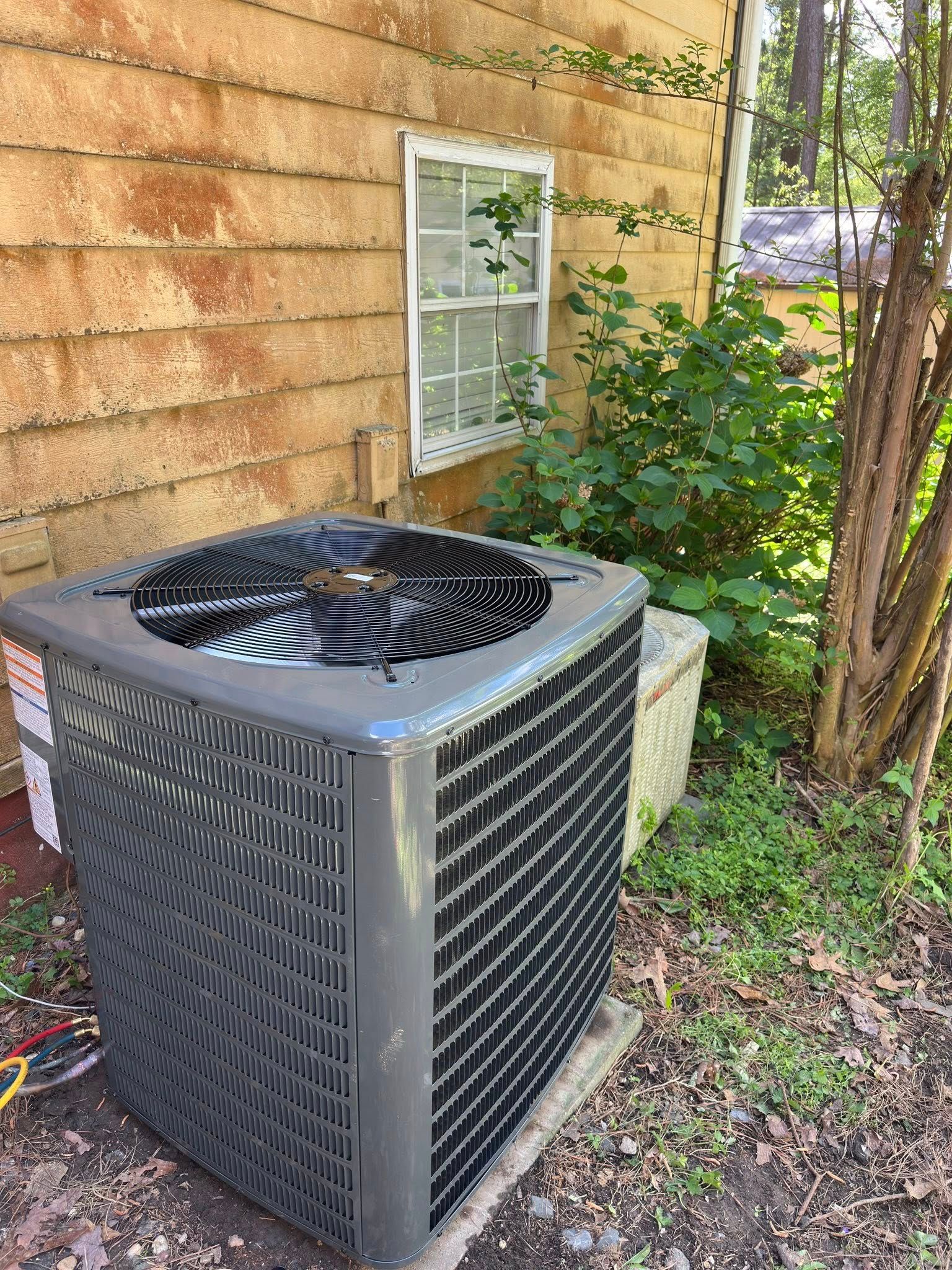 Air conditioning unit next to a light-colored building, small window, and greenery.