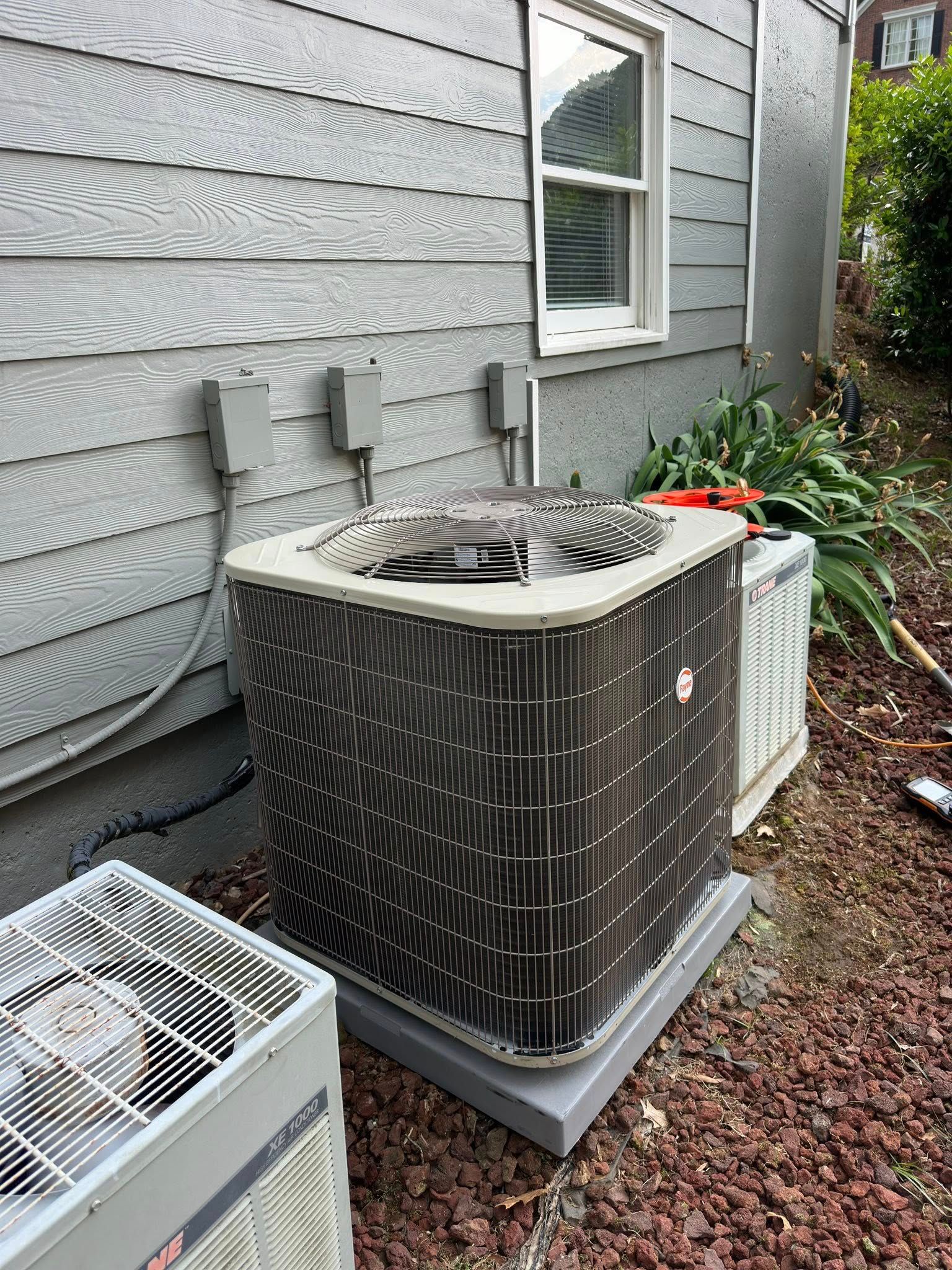 Two AC units outside a building with gray siding, next to a window and bushes.