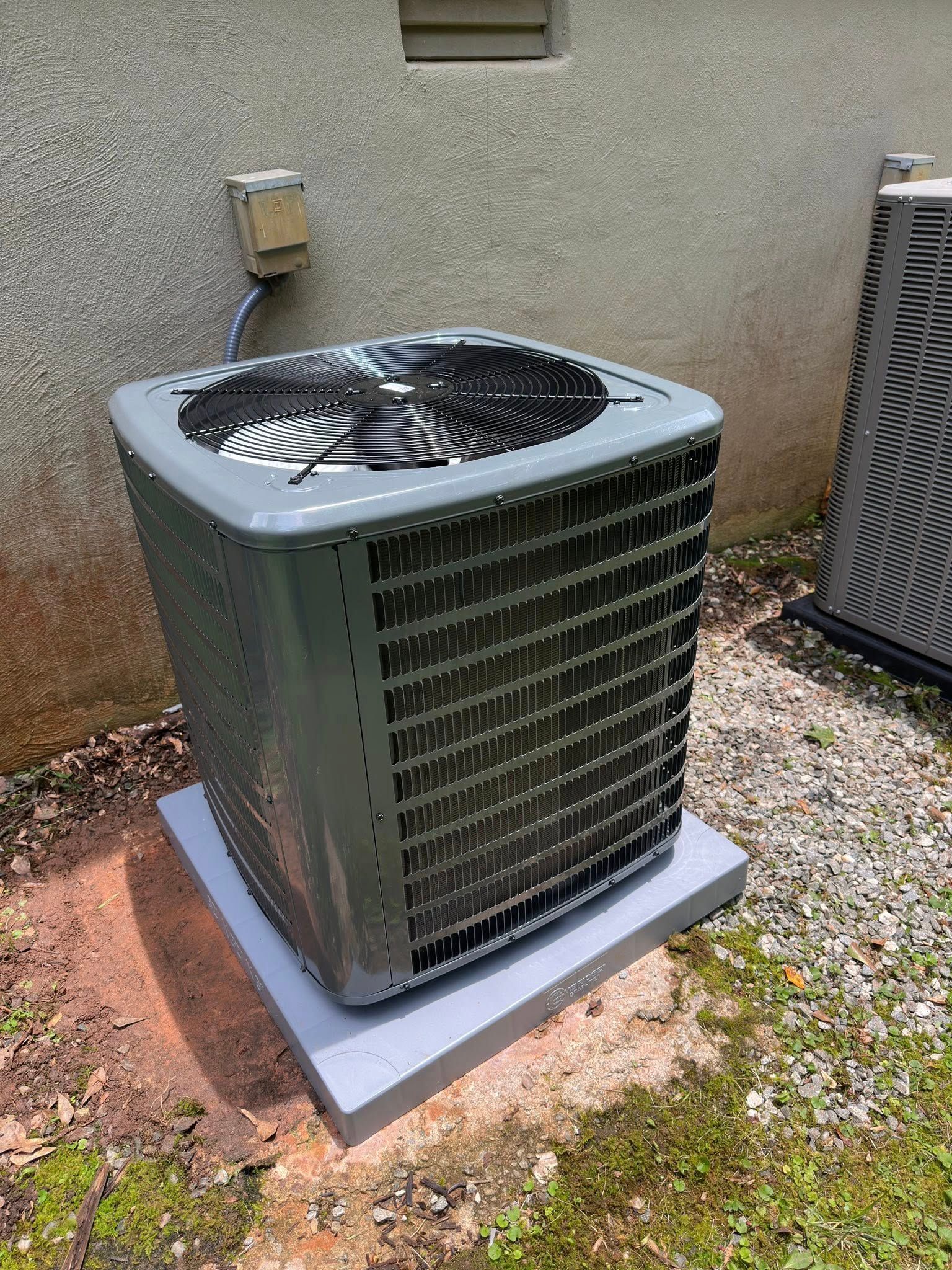 Outdoor air conditioning unit on a concrete pad near a building. Gray and silver with a black fan.