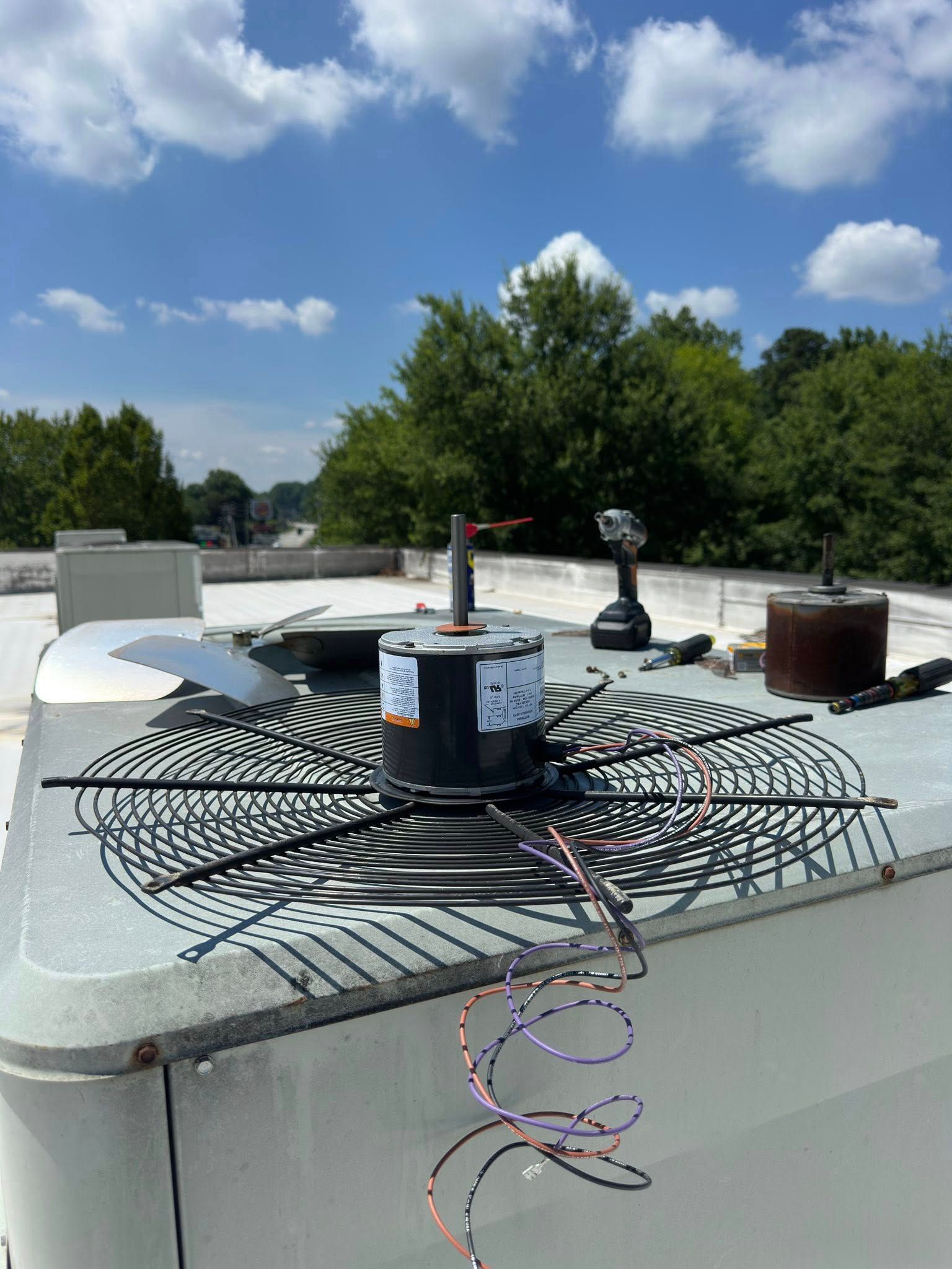 An HVAC unit on a rooftop with motor replacement underway; blue sky, trees in background.