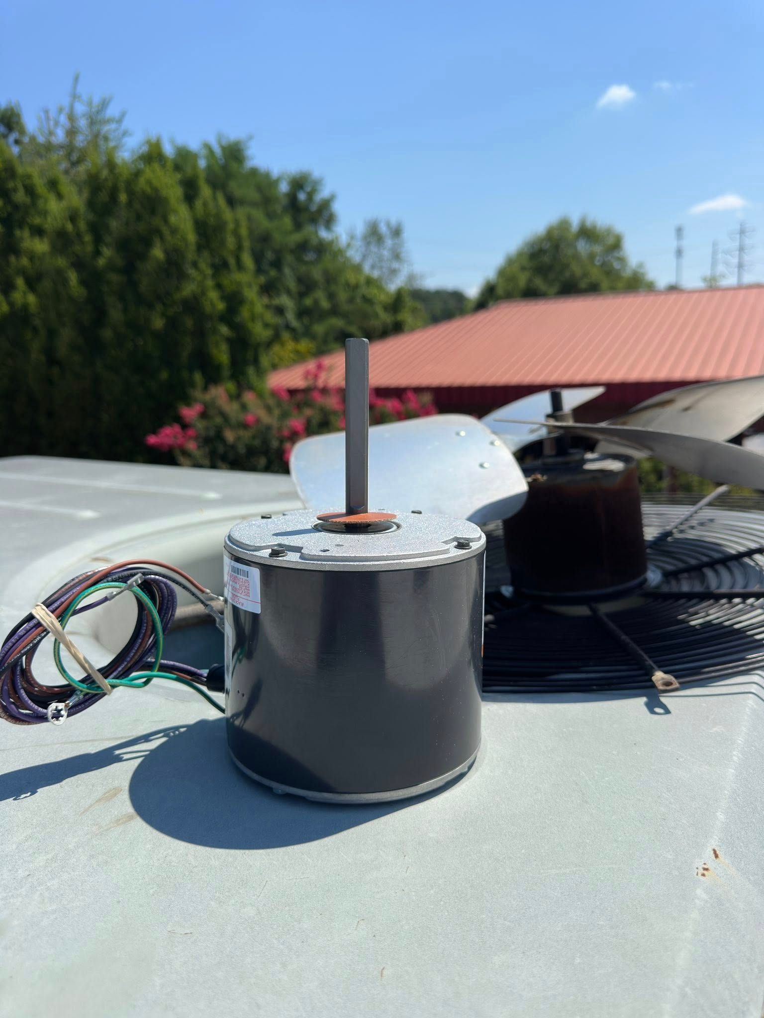 AC condenser fan motor with attached wires, sitting on a rooftop with a sunny, clear sky in the background.