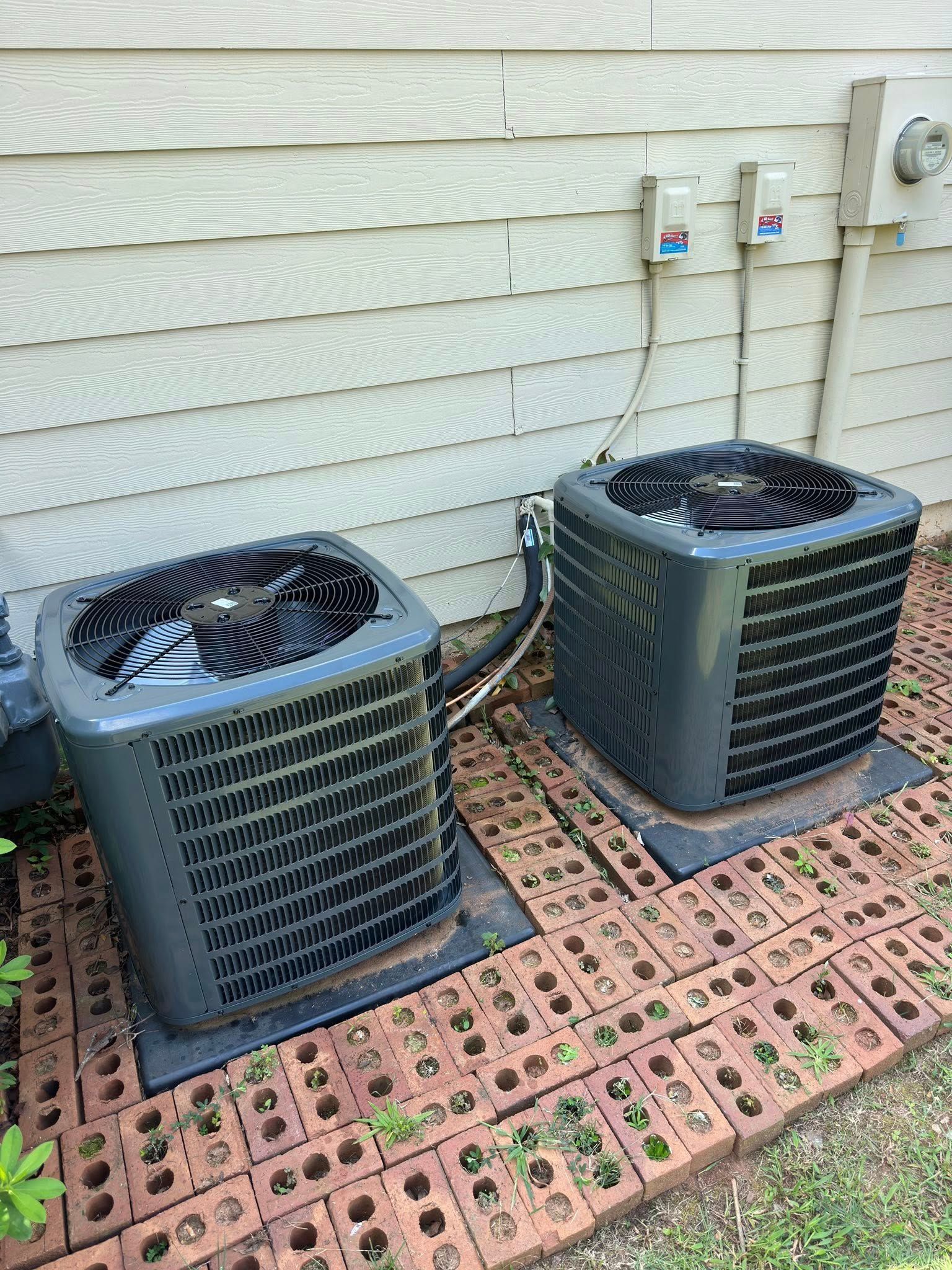 Two gray air conditioning units sitting on brick pavers near a house.