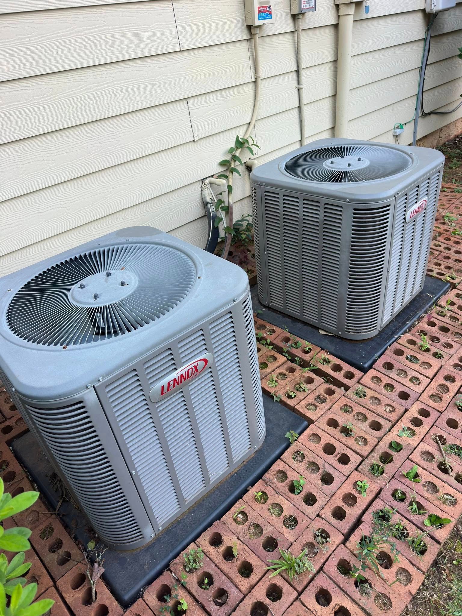 Two gray Lennox air conditioning units on black pads, brick patio, against a beige siding wall.