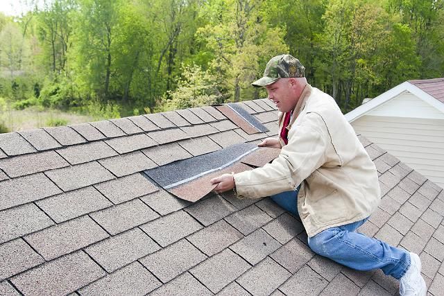 Man on rooftop repairs asphalt shingles; green trees in background.