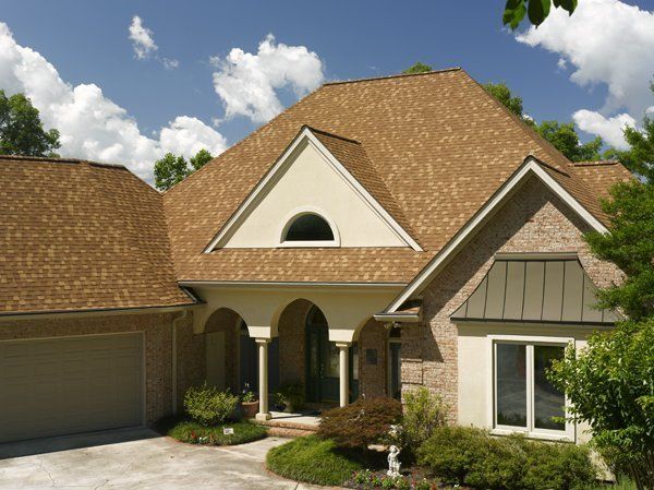 Brown-roofed house with beige siding, arched entryway, and garage. Blue sky with clouds.