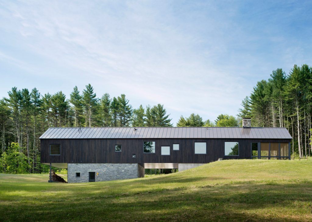Modern black wooden house with large windows, on a grassy hill, backed by trees, under blue sky.