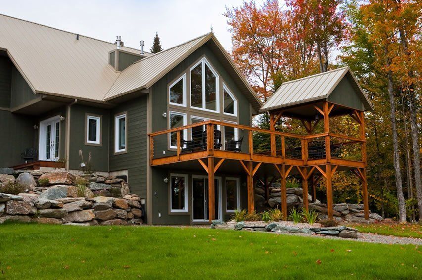 Green house with large windows, wooden deck, and tan metal roof, surrounded by trees with fall foliage.