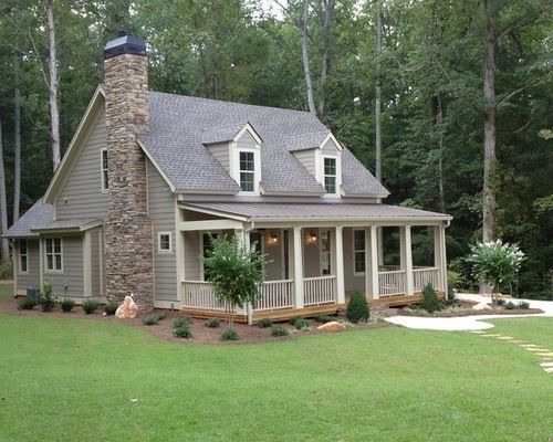 Cottage with stone chimney, wraparound porch, and gray siding surrounded by trees on a green lawn.