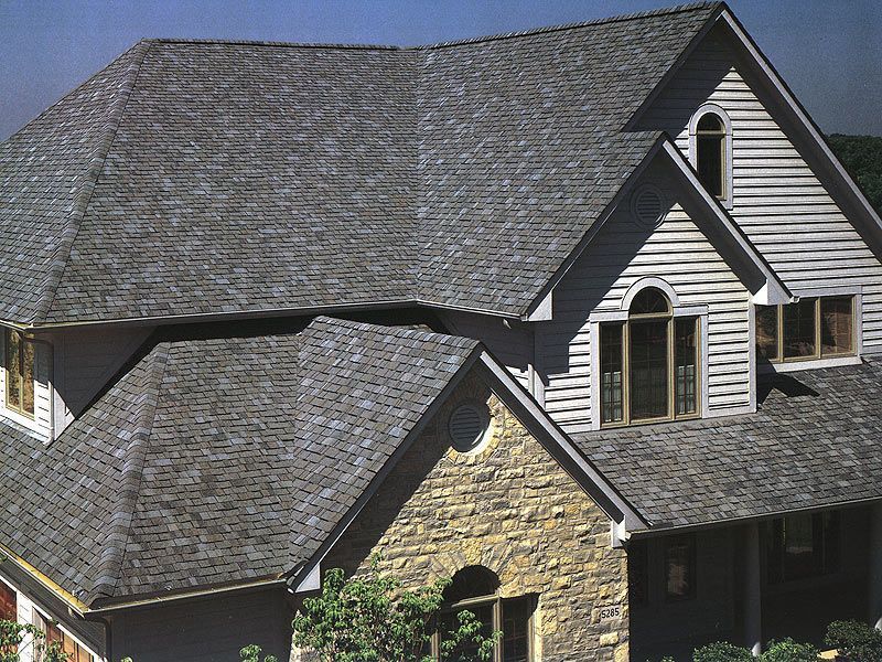 House with gray shingle roof, beige stone and light siding, and arched windows.