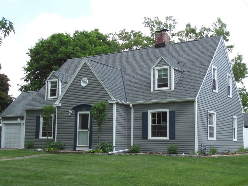 Gray Cape Cod-style house with blue shutters, green lawn, and trees.