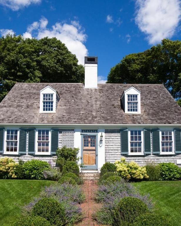 Cottage with gray shingle siding, green shutters, and a walkway through a garden, under a blue sky.
