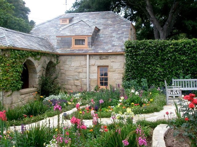 Stone building with slate roof, arched doorway, and colorful flower garden. Bench on right.