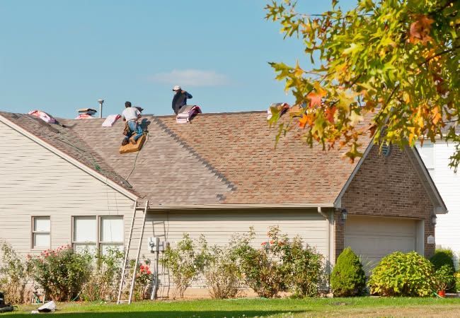 Roofers replacing shingles on a house roof under a sunny sky.