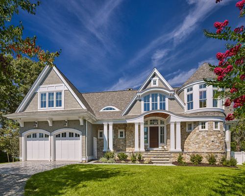 Gray and stone-sided house with a lawn, white garage doors, and a blue sky.