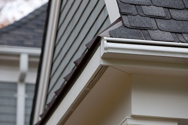 White gutter along the edge of a house roof, with gray shingles and siding visible.