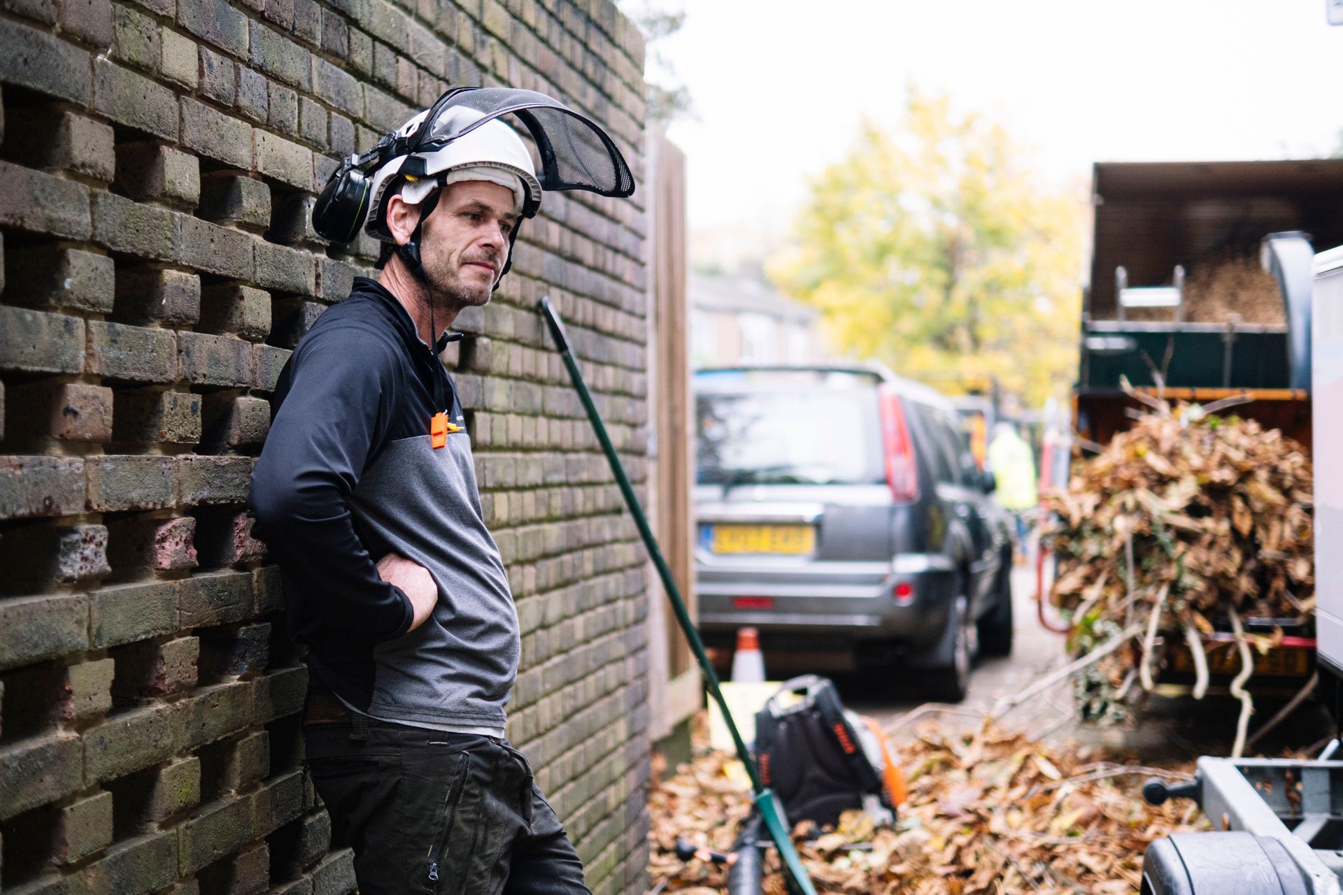 Man in safety gear leans against a brick wall, watching leaves being loaded into a truck.