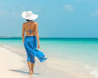 A woman wearing a blue sarong and a white hat is walking on the beach.