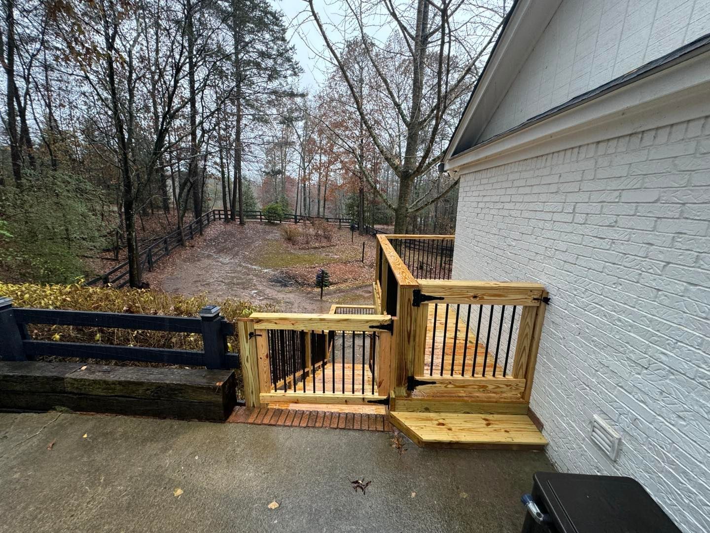 A wooden deck staircase and safety gate with black metal balusters attached to a white brick house near a wooded yard.