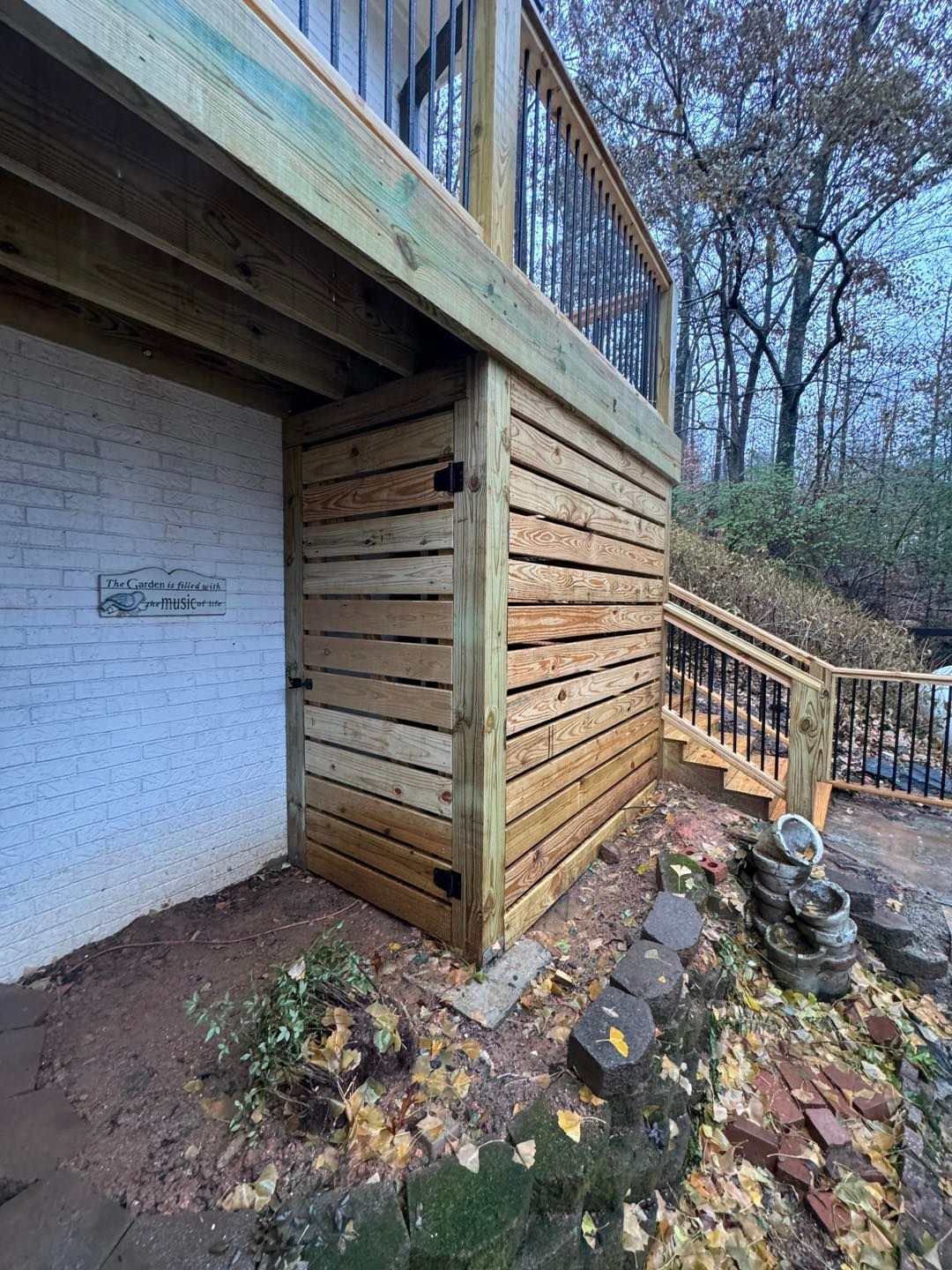 A wooden under-deck storage enclosure with a hinged door, positioned beside a white brick wall and stairs.