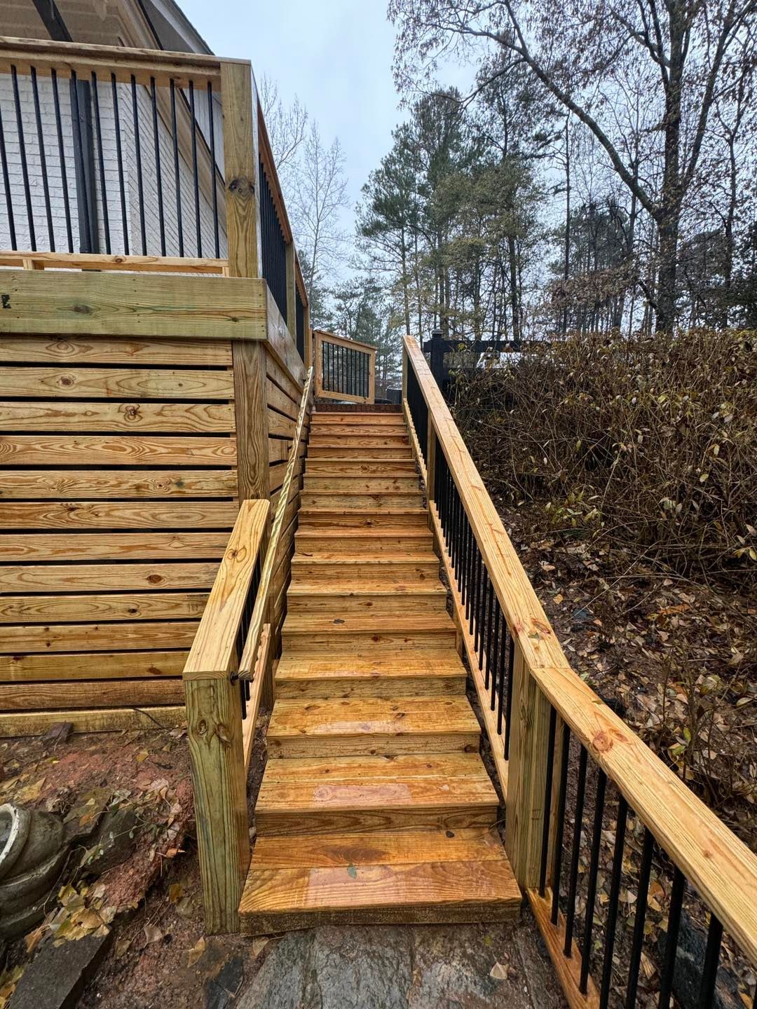 A long set of wooden outdoor stairs with black metal railings leading up to a deck, set against a wooded hillside.