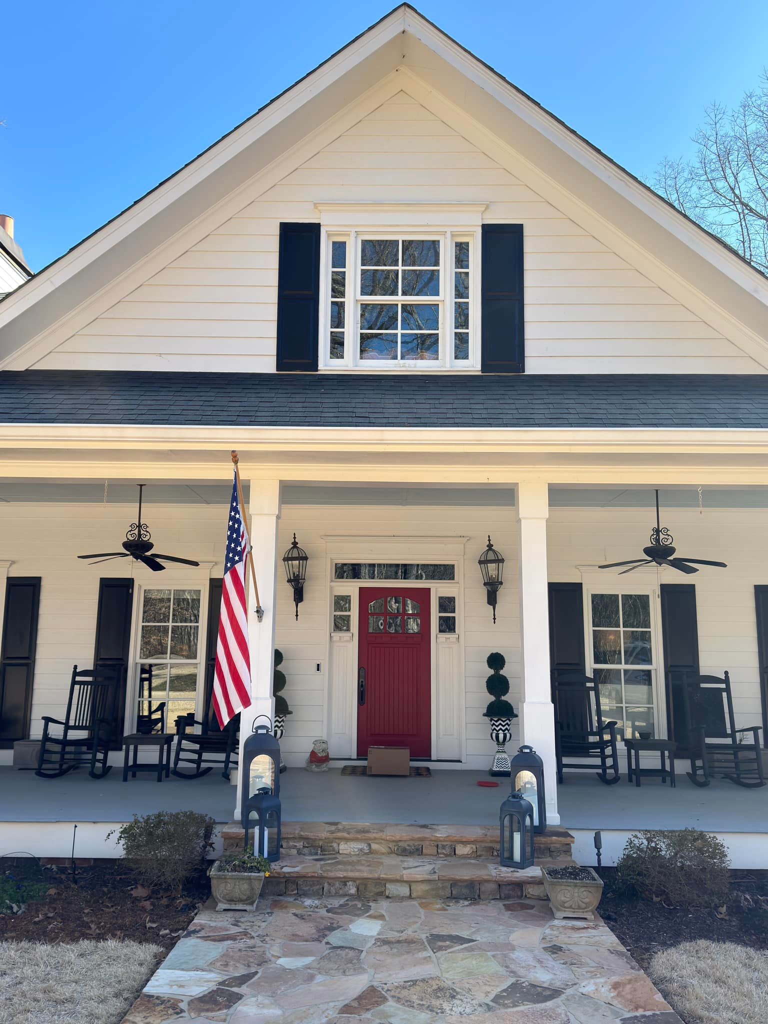 A two-story white farmhouse with a stone porch, a red front door, an American flag, and black shutters under a blue sky.