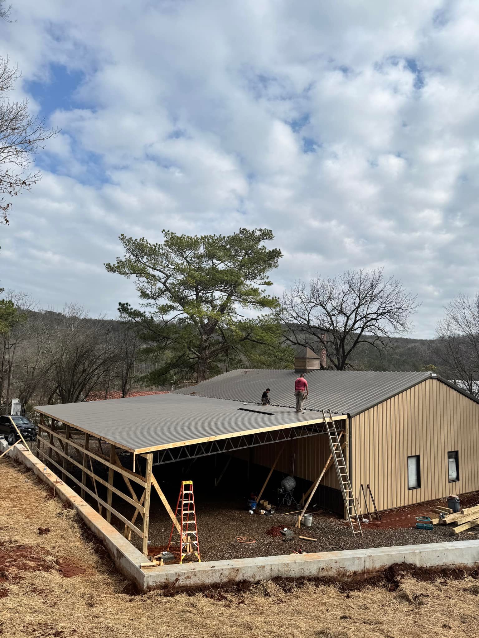 Workers install metal roofing on a tan, partially finished pole barn building on a rural site.