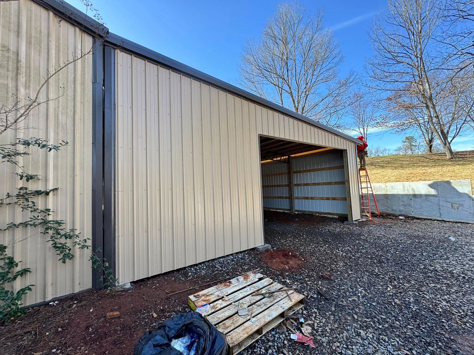 A person stands on a ladder near the entrance of a beige metal building with a gravel foreground.