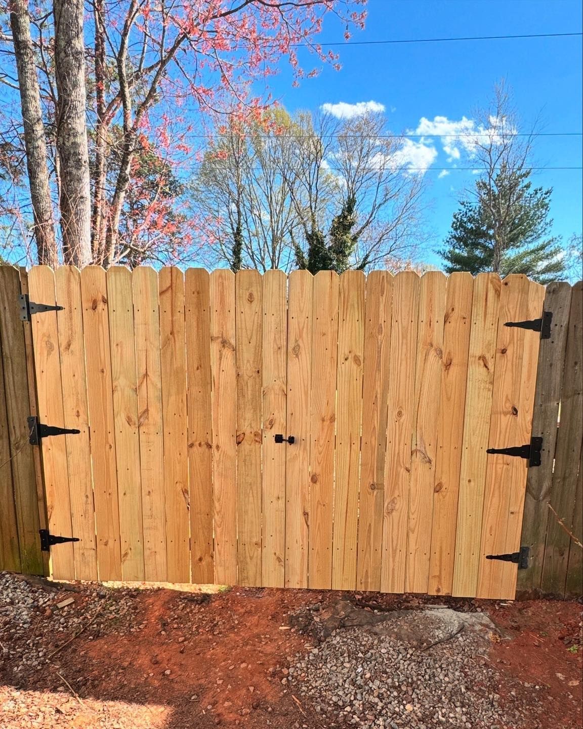 A wooden double-gate fence with black hinges and a center latch, set against a background of trees and a blue sky.