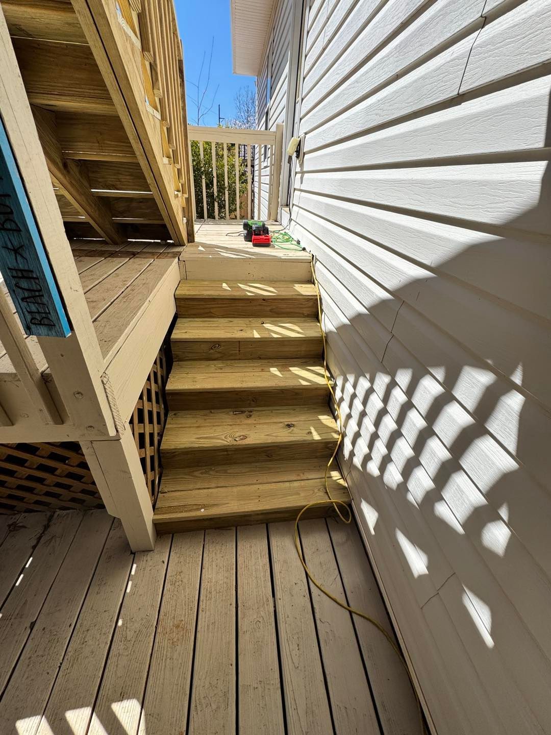 Wooden stairs leading up to an outdoor deck landing next to white vinyl siding on a sunny day.