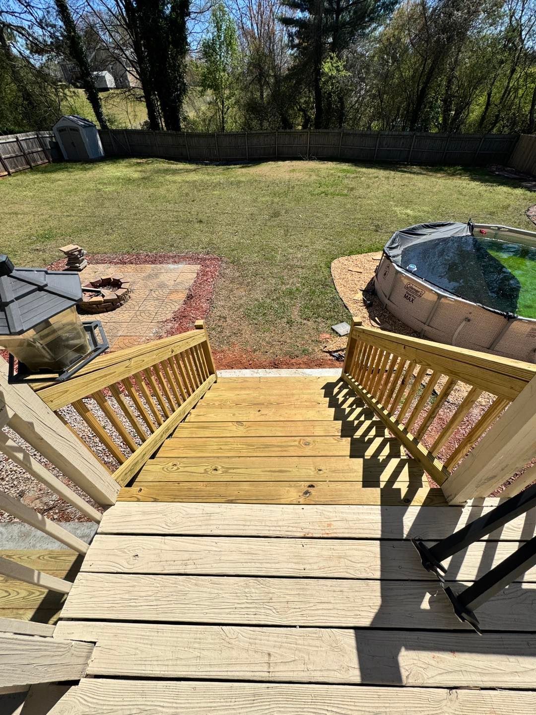 A backyard view from a wooden deck, overlooking a lawn with a small fire pit area on the left and an above-ground pool.