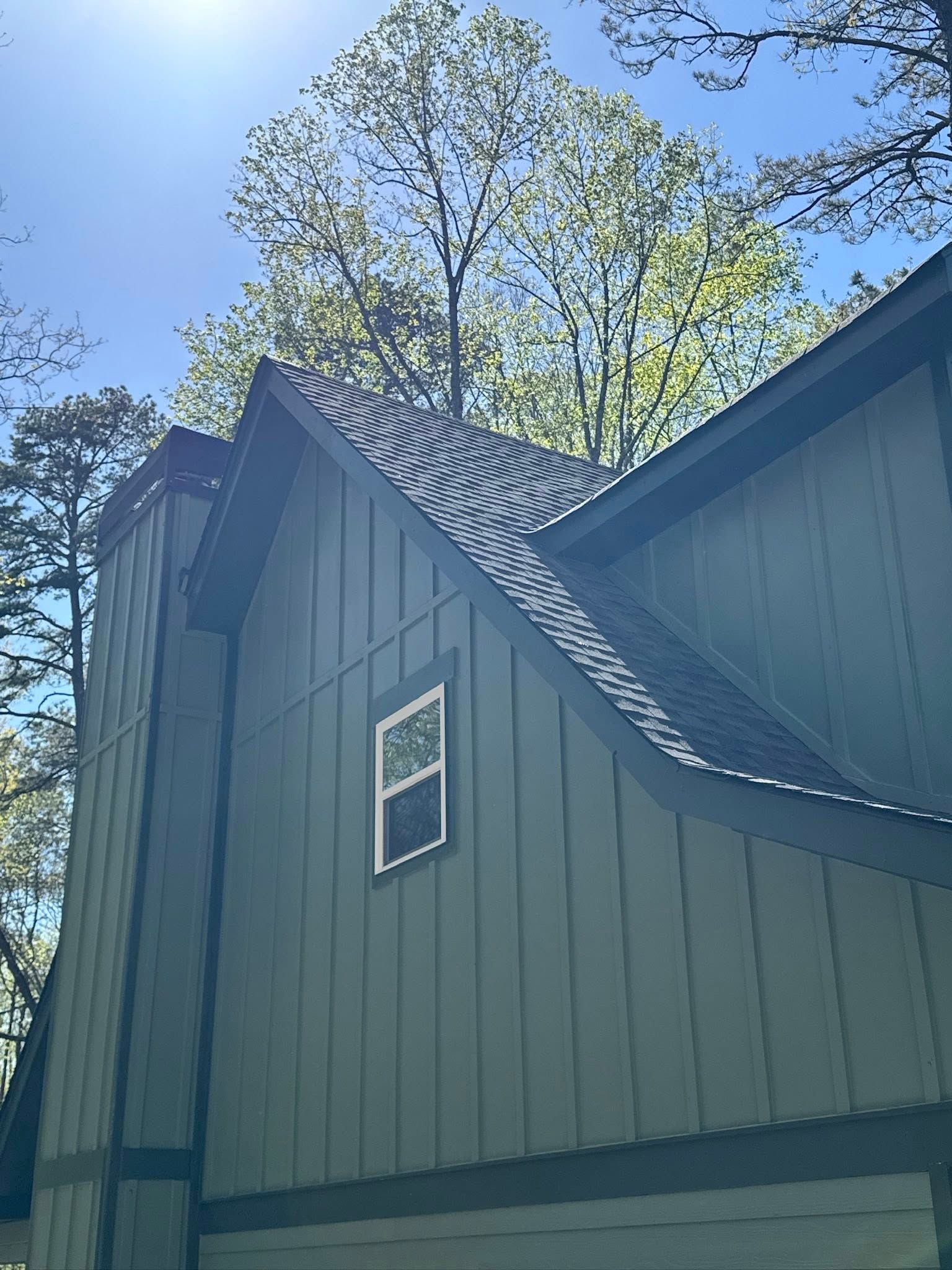 A low-angle view of a sage green, board-and-batten house exterior with a dark roof and a small, rectangular upper window.