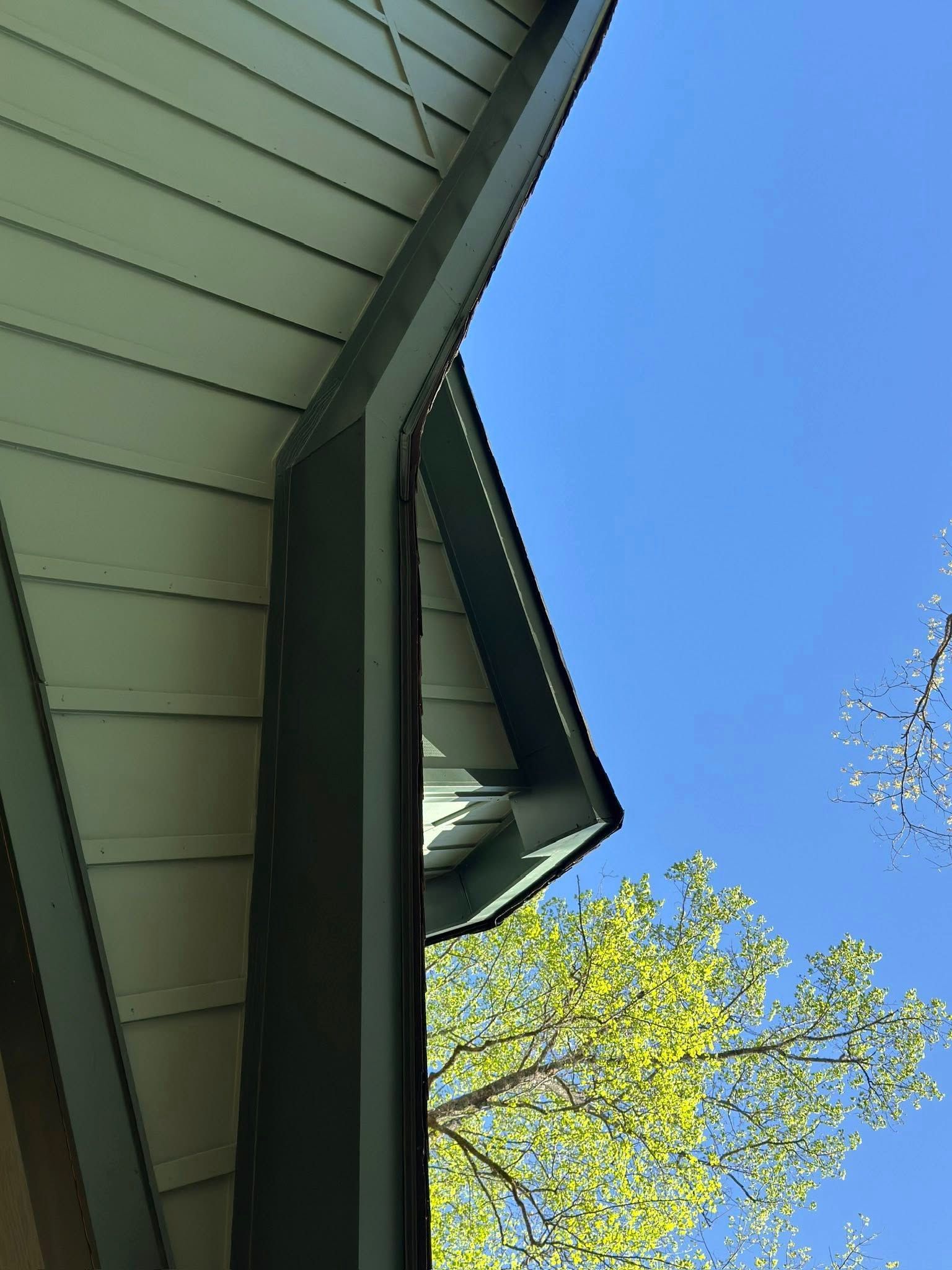 A low-angle view of a house corner showing beige siding, dark trim, and a downspout against a clear blue sky.