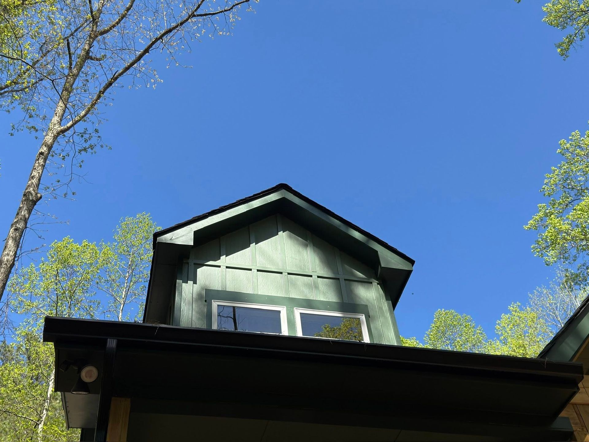 A green cabin dormer with windows, surrounded by trees against a clear blue sky.