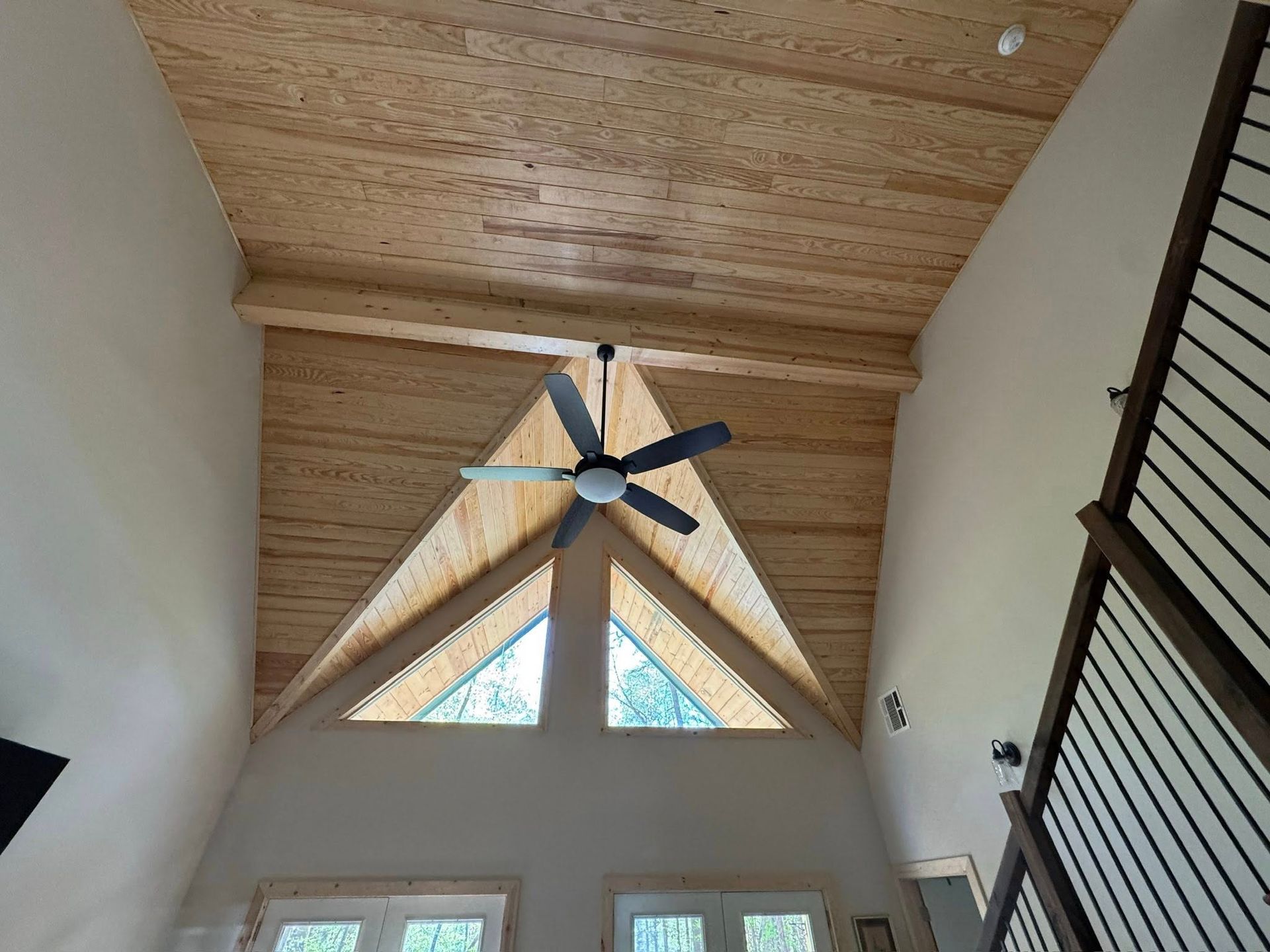 A vaulted room with a wood-plank ceiling, exposed beam, ceiling fan, and triangular windows above glass doors.