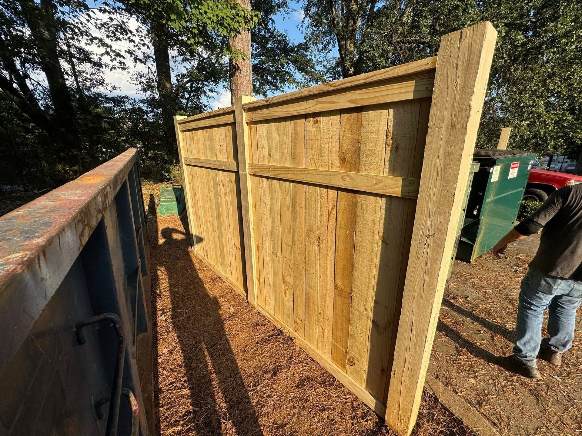 A newly installed wooden fence section stands outdoors next to a large metal dumpster, with a person standing nearby.