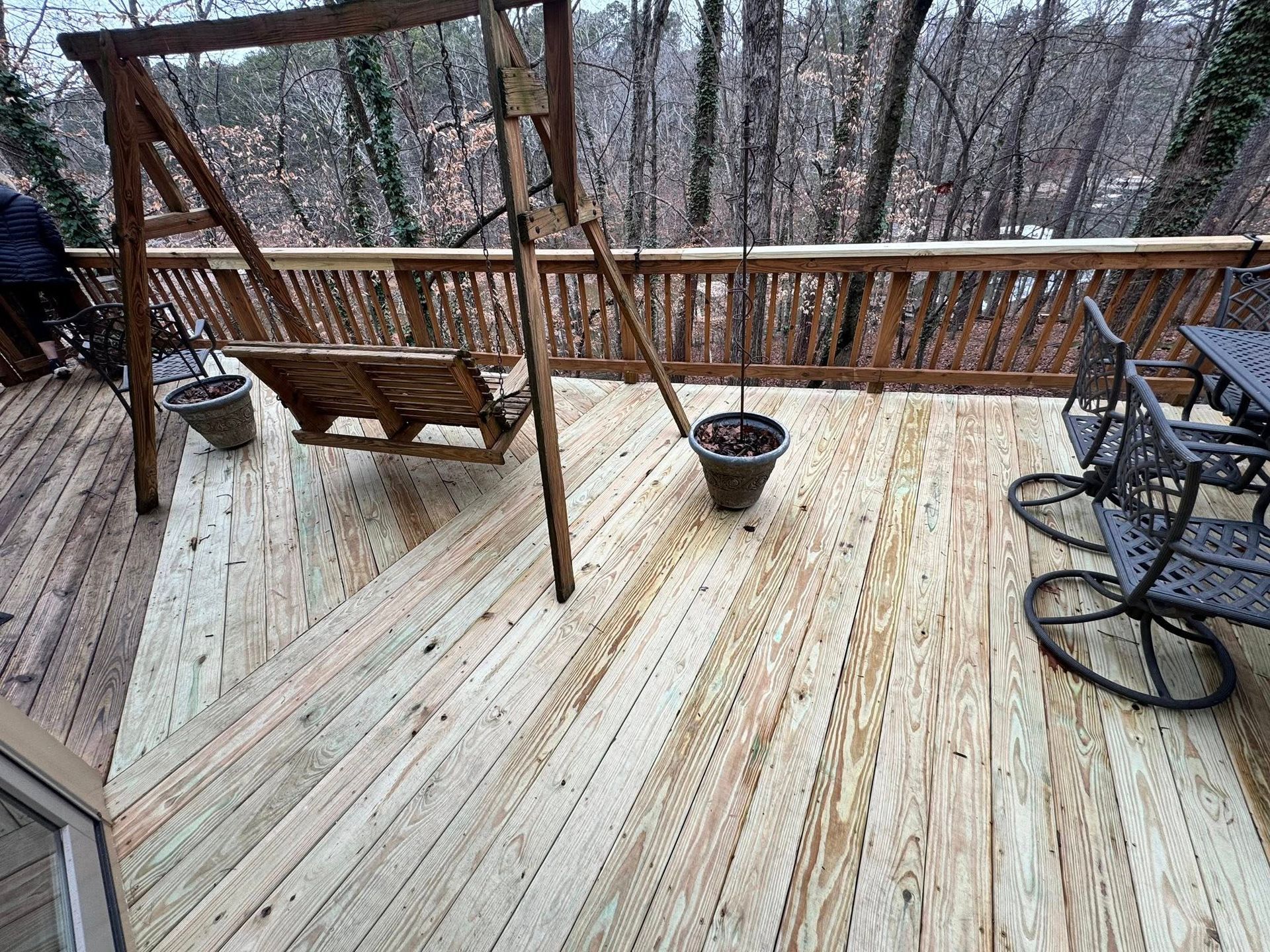 A wooden deck with a swing bench, a potted plant, and metal patio chairs overlooking a forest during a winter day.