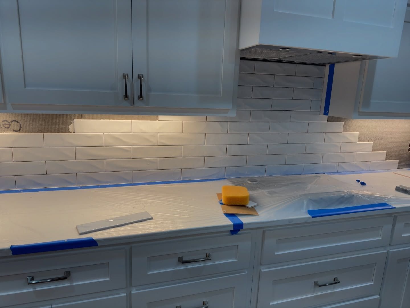 White subway tiles being installed on a kitchen backsplash, with blue painter's tape on the white countertops and cabinets.