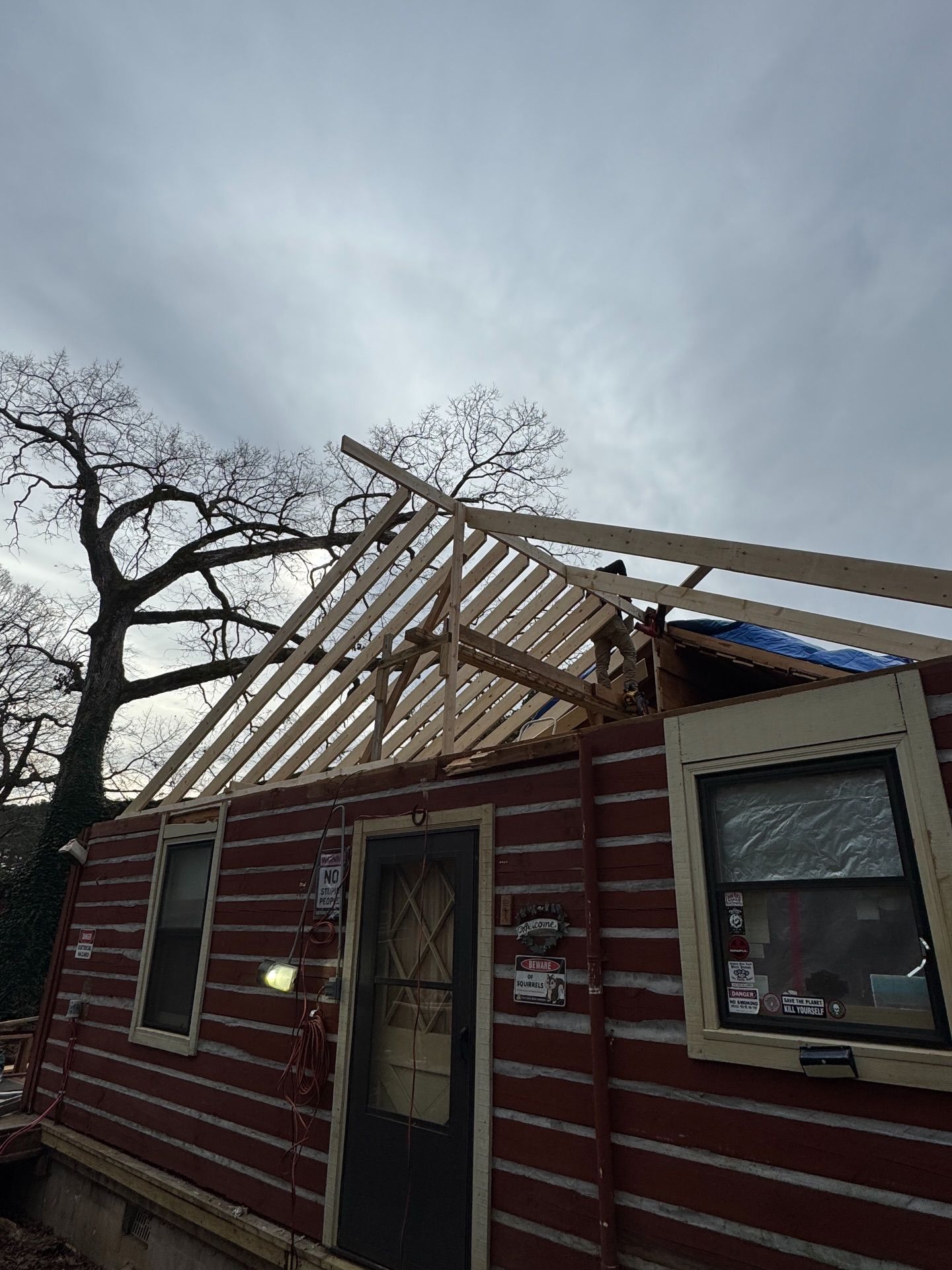 A low-angle view of a red-sided house under renovation, showing exposed wooden roof rafters against a cloudy sky.