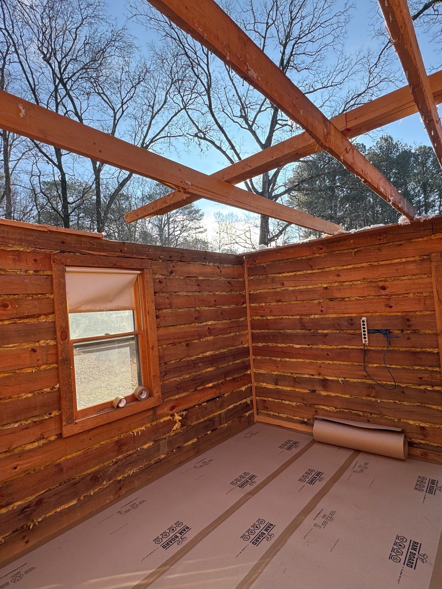 Interior view of a rustic log cabin under construction with exposed wooden ceiling beams and flooring underlayment.