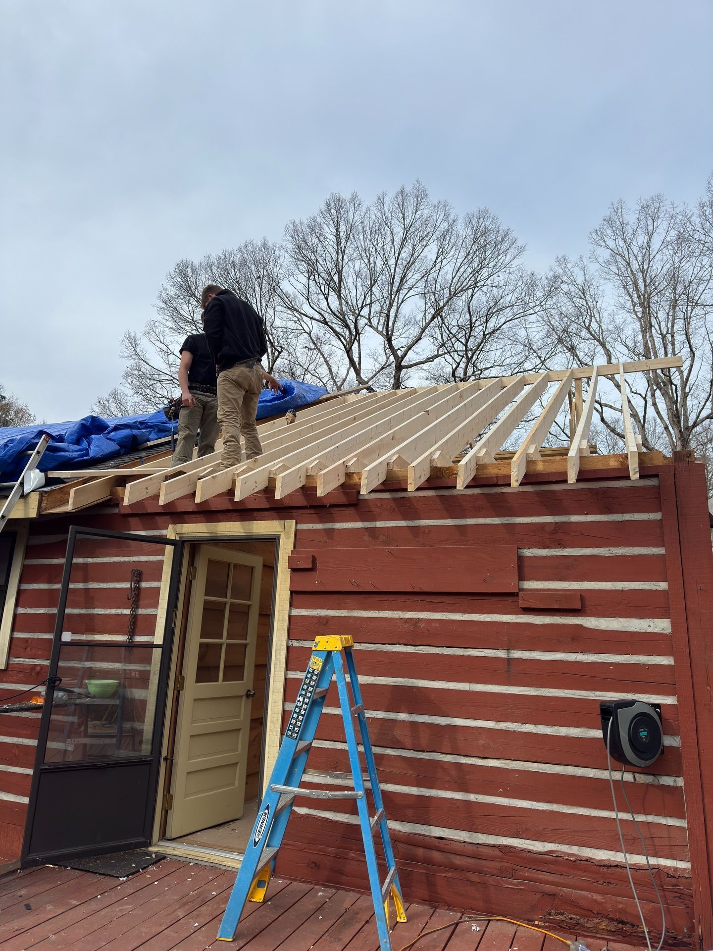 Two people work on the wooden rafters of a red log cabin roof, with a blue ladder standing on a deck below.