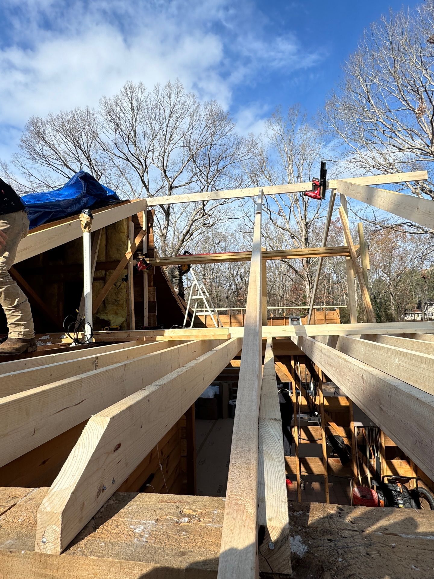 Construction workers frame a wooden roof structure with new beams against a bright blue sky and bare trees.